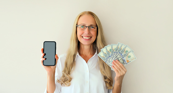 Portrait of happy smiling middle-aged woman holding blank screen phone and cash money in dollar bills on white studio background