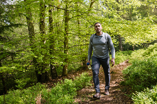 A wide angle full length view of a young man carrying a rucksack wearing athletic leisure wear and enjoying a hike in the woodland in Northumberland in the North East of England. He is also carrying his water bottle to keep hydrated.
