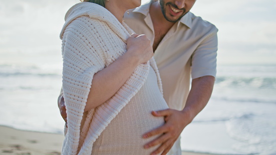 Happy man touching pregnant belly beloved wife walking sunshine beach closeup. Married couple relaxing together on summer seashore expecting baby. Young future parents enjoy romantic vacation laughing