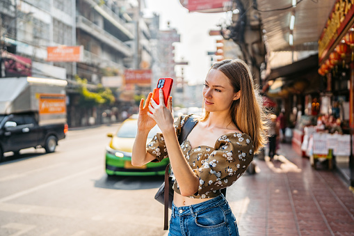 Beautiful young woman taking pictures using her smart phone on the street in Chinatown in Bangkok in Thailand.