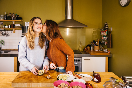 Young female couple kissing while preparing breakfast in their home kitchen. A lesbian couple shares an intimate moment together, savoring tea and exchanging kisses in the cozy atmosphere of their kitchen at home.