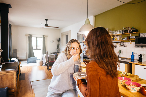 A lesbian couple eating breakfast and sharing an intimate moment together, in the cozy atmosphere of their kitchen at home.
