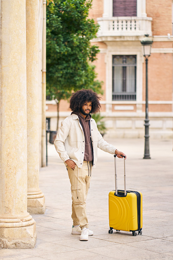 A man wearing a white jacket and tan pants is holding a yellow suitcase. He is standing in front of a building with a tree in the background. Concept of travel and adventure