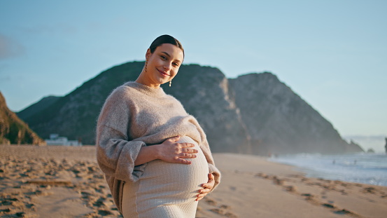 Pregnant girl posing seashore with happy smile. Beautiful expectant mother looking camera standing on sand beach enjoying pregnancy. Dreamy woman expecting baby contemplating ocean waves at evening.