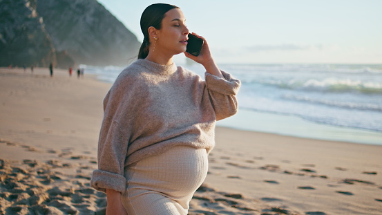 Pregnant girl calling at beach caressing big belly. Happy carefree woman expecting baby talking on smartphone walking beautiful sand coast. Tender future mother smiling enjoy phone communication.