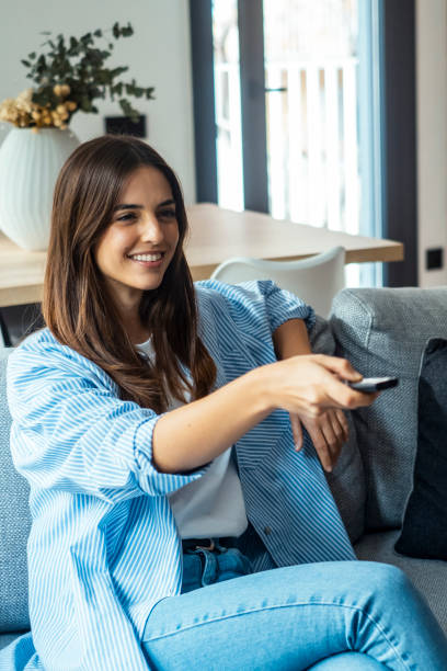 istock young woman sitting on her home sofa, holding the TV remote while enjoying a program. she is enjoying her leisure time at home. One female people using control tv watching movie alone on the couch 2085884747