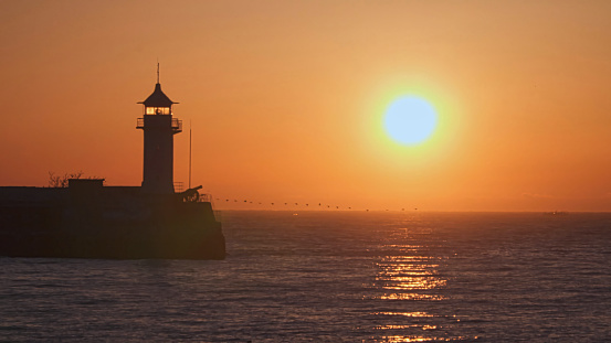 The lighthouse in the morning. There is an ancient cannon near the lighthouse. Sunrise at sea. There is a flock of birds on the horizon