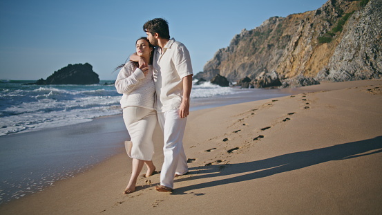 Couple awaiting child walking sandy shore at summer family vacation. Relaxed romantic husband hugging pregnant wife on sunny sea coast going near ocean waves. Carefree future parents enjoy pregnancy.