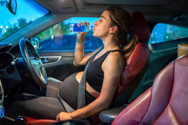 istock Side view pregnant woman sitting in car and drinking water from plastic bottle while waiting traffic light to change. 2085987032