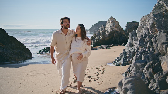 Expectant father mother walking beautiful rocky shore hugging together. Carefree romantic pregnant couple stepping sand near rough stones seacoast. Happy married pair enjoy summer vacation on ocean.