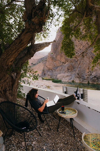 Side view of Middle Eastern female sitting at the terrace with canyon view remote working and discussing job issues by laptop in the mountains of Oman