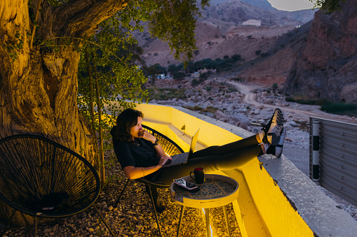 Side view of Middle Eastern female sitting at the terrace with canyon view remote working and discussing job issues by laptop in the mountains of Oman