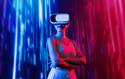 Smart female standing surrounded by neon light wearing VR headset connecting metaverse, future cyberspace community technology. Elegant woman confidently pose crossed arm look faraway. Hallucination.