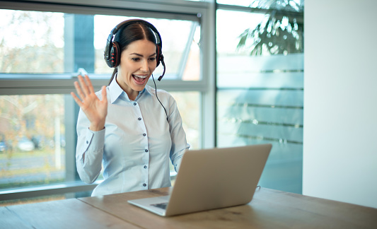Smiling businesswoman in headset waving hand while making video call with customer at workplace. Female call center service support agent talking to a client during online meeting.