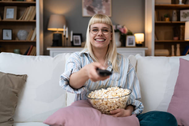 istock Young happy woman changing channels with remote control while watching TV and eating popcorn in the evening at home. Relaxed hipster female enjoying at weekend lazy day 2086091559