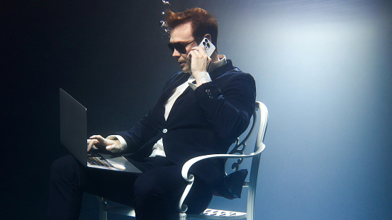 Businessman in a suit is sitting underwater with a laptop and talking on a mobile phone