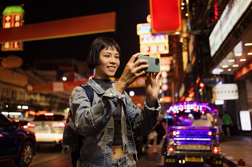 Happy Asian woman taking a photo with her mobile phone on the street at night.