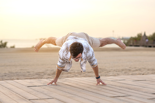 Handsome man in casual clothes doing handstand on his finger at sidewalk path at beach during sunset