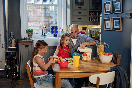 A father and his two daughters sitting at the kitchen table in the kitchen at home in South Shields, North East England. They are eating and enjoying breakfast together and the man is pouring milk on his daughter's cereal as she watches.