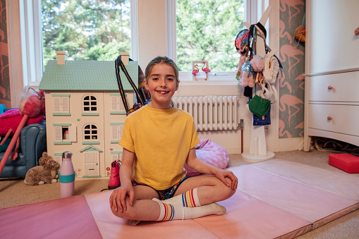 A young girl sitting on an exercise mat in her bedroom at home in South Shields, North East England. She is taking a break from meditating while looking at the camera and smiling.
