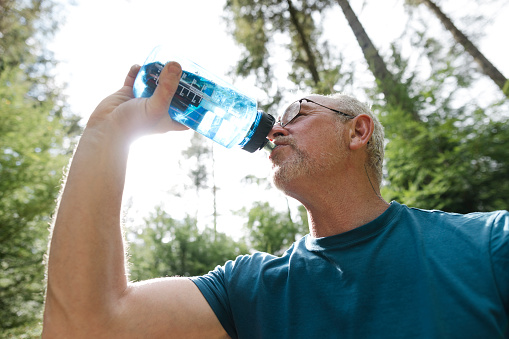 A low angle close up of a mature man stood outdoors in a forest in the North East of England. He is using a reusable bottle and stands having a mindful moment and appreciating the green surroundings.