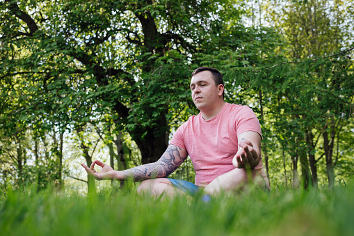 A medium close-up of a young man who is sitting meditating in a lush green forest area in the North East of England. He is enjoying the ambient forrest noises as she enjoys the surroundings and feeling connected to nature.