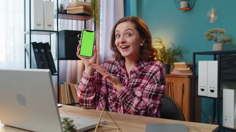 Woman freelancer looking at camera and showing smartphone with green screen for new application