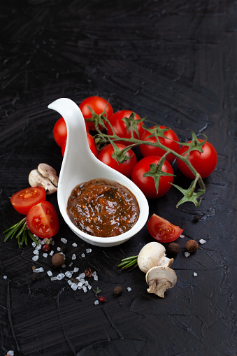 A marinade for shashlik in a white cup, set against a black background, accompanied by spices and red tomatoes on a branch