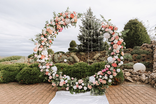 Modern ceremony in European style. Wedding arch with roses on the background of a decorative garden. Jewelry from fresh flowers, flowers and crystals. Front view.