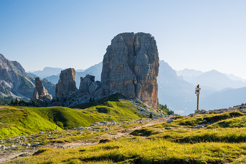 Cinque Torri peaks at summer cloudless morning with green grass lighteen by warm light of sun and guidepost against rocky mountains