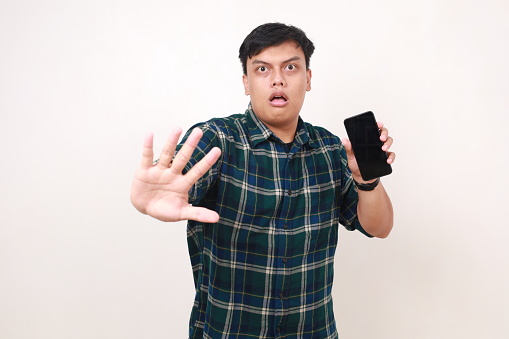 Serious young asian man with stop hand gesture while showing a blank phone screen