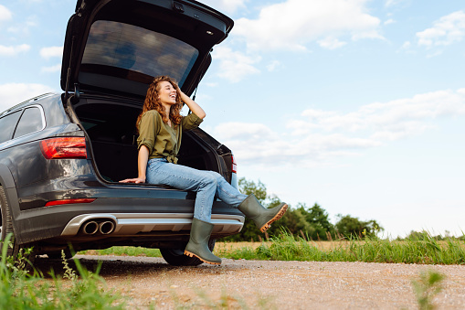 Young woman sitting in open trunk, resting and enjoying journey. Lifestyle, travel, tourism, active life.