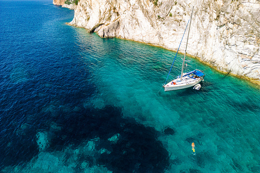 A sailboat anchored in a beautiful bay of Meganisi island in Greece. The sea is crystal clear, blue and turquoise and cove is surrounded with rock cliff. One person snorkeling near boat