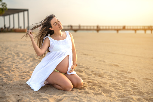 Pregnant woman wearing white dress sitting over sea shore outdoors.