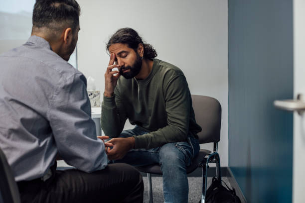 Feeling Sad at Hospital Over-the-shoulder shot of a doctor and man. The man looks sad and depressed after hearing news from the doctor. The man's eyes are closed and has one hand covering part of his face. edgy man stock pictures, royalty-free photos & images