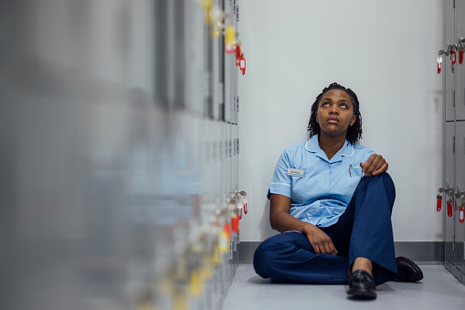 Medium shot of Nurse sitting down in a locker room. She is sat with one leg up and her arm resting on it. The woman is looking up with her back against the wall. She is wearing a blue nurse uniform.