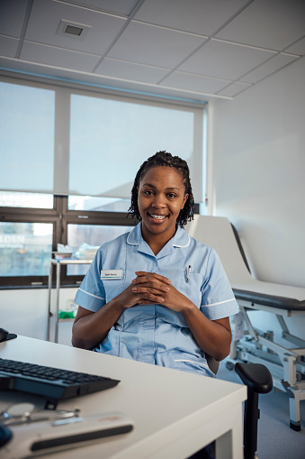 medium shot of a nurse sitting at a desk. She is sitting at a desk in an office chair. The woman has both hands intertwined. She is looking at the camera with a. smile on her face. The nurse is wearing a blue nurse uniform.