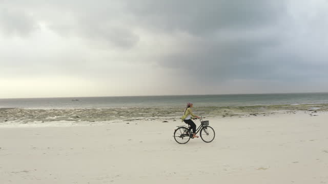 4K footage of young woman riding on bicycle along beautiful packed damp sand beach. Spending time outside, sports, leisure activities concept.. Environment, healthy lifestyle, outdoors concept.