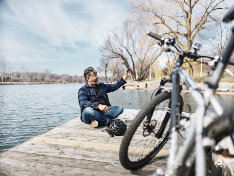 Mature Indian man taking a selfie while resting from  cycling on the lake pier. Dressed in casual clothes. Exterior of parking lot in public park.