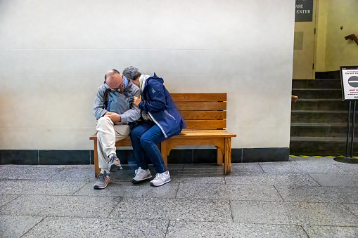 Ellis Island, New York, NY, USA – September 21th, 2023: A middle-aged man and woman wearing sneakers hold each other while sleeping on a bench in the buildings on Ellis Island New York. The man has a backpack on his lap.