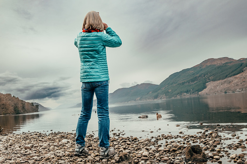 Senior woman on the shores of Loch Ness in Scotland, a place where there is said to be a monster hiding in the deep waters. Best to carry a pair of binoculars, just in case.