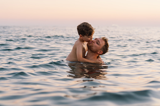 A father and son share a pleasant moment while swimming in the calm ocean waters as the evening light gently fades in the background. Their smiles convey a sense of enjoyment. Happy Fathers Day