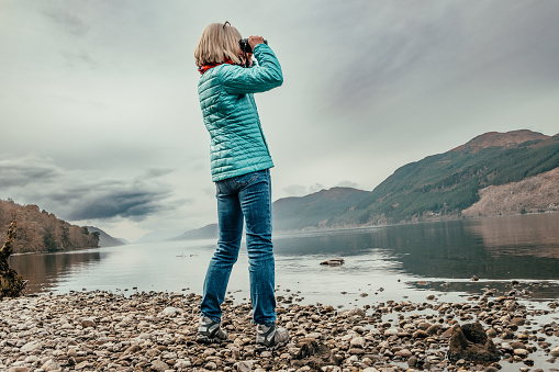 Senior woman on the shores of Loch Ness in Scotland, a place where there is said to be a monster hiding in the deep waters. Best to carry a pair of binoculars, just in case.