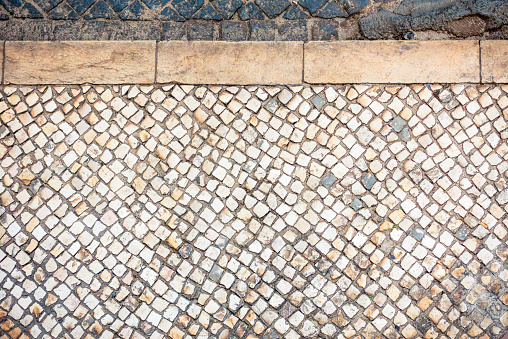 A view of an old and heavily weathered sidewalk of mosaic paving stones, and a traditional stone curb, photographed from directly above in Lisbon, Portugal.