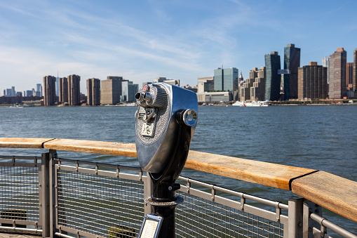 Queens, NY, USA–September 21th, 2023: Close-up of silver public binoculars on the pier of Gantry Plaza State Park overlooking the East River and the skyscrapers of midtown Manhattan.