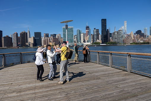 Queens, NY, USA–September 21th, 2023: A group of tourists take photos of Manhattan from the pier at Gantry Plaza State Park. A man in a yellow shirt helps Asian tourists with their camera.