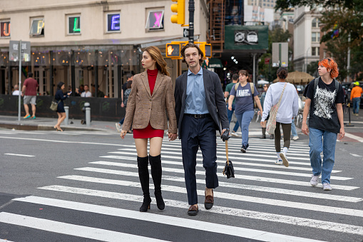 Manhattan, NY, USA–September 22th, 2023: A young man and woman walk across a crosswalk in the West Village. The woman is wearing a short skirt with a blazer and high boots. The man in a suit carries her handbag.