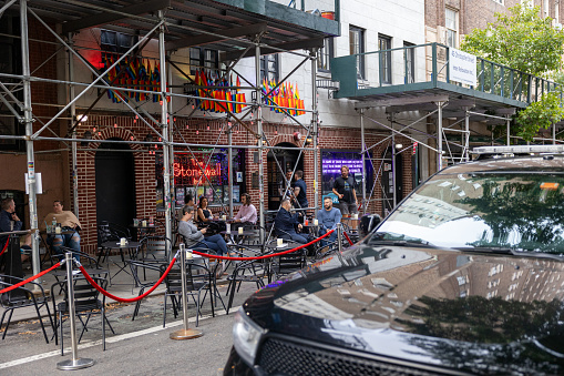 Manhattan, NY, USA–September 22th, 2023: Street scene of men and women sitting on the terrace  of the scaffolded Stonewall Inn in Christopher Street West Village. There is a black car in front of it.