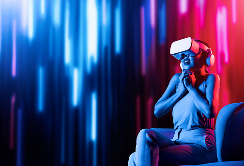 Smart female sitting on sofa surrounded by neon light wearing VR headset connecting metaverse, future cyberspace community technology. Elegant woman excited and emotionally watch movie. Hallucination.