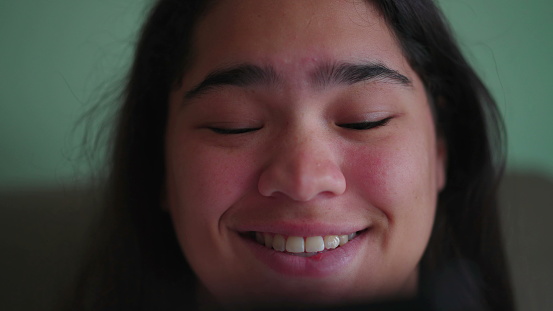 Joyful Brazilian teen girl react_s positively to content online while holding phone. Close-up of a happy diverse Asian woman_s face smiling reading message